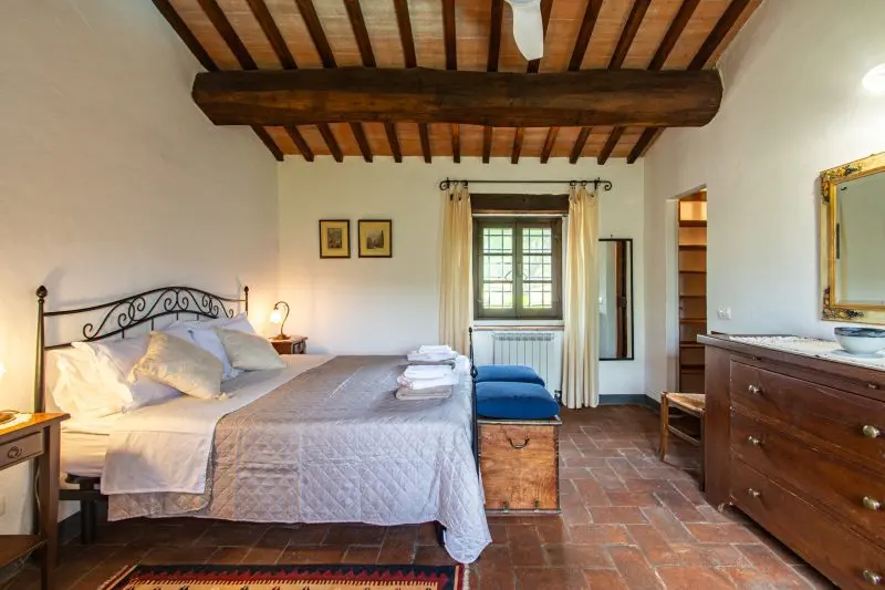 Rustic master bedroom with exposed wooden ceiling beams, wrought-iron bed, and cream window curtains.