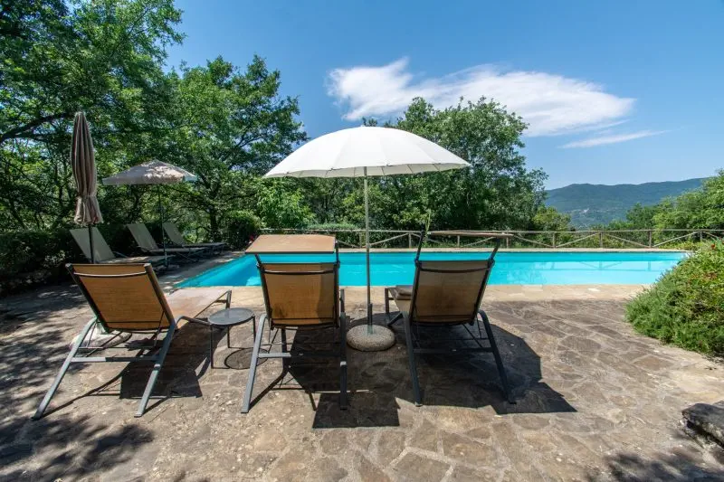 Poolside area with brown lounge chairs and a large white umbrella on a stone patio, surrounded by trees and distant hills under a blue sky.