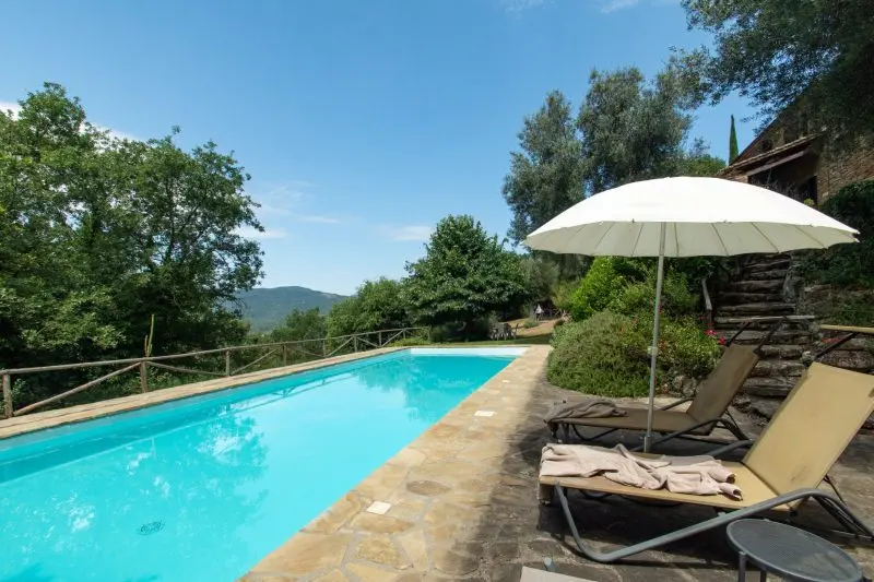 Poolside scene with a bright blue rectangular pool, stone patio, lounge chairs, and a large white umbrella among green trees and a stone building nearby.