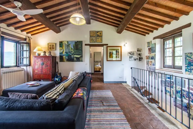 Cozy rustic living room with exposed wooden beams, a dark sectional sofa, and colorful cushions under a sloped ceiling. West-facing windows brighten the space.